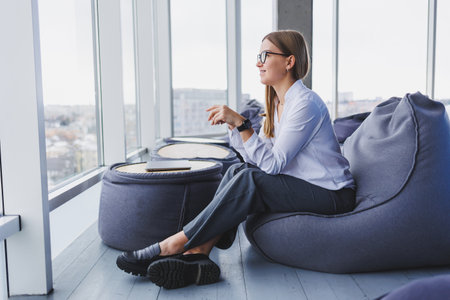 Relaxed smiling happy young woman in glasses and white shirt sitting in bean bag chair, resting, using laptop work, making notes in notepad, computer, remote work conceptの写真素材