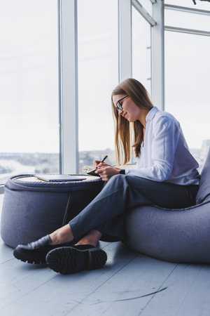 Break. Smiling young woman in glasses relaxing on bean bag chair sitting at table and relaxing using notepad, happy woman leaning back enjoying work feeling satisfiedの写真素材