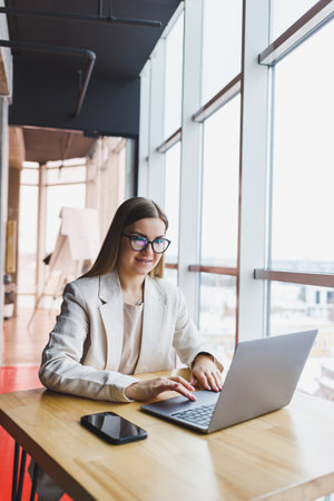 Focused woman in casual wear typing on a netbook while working on a new project, sitting at a desk in a modern workspace. Business lady in a jacket and glasses. Remote workの写真素材