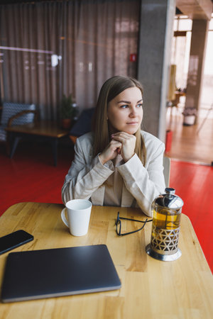 A beautiful woman of Slovenian appearance, a manager in a light jacket and glasses, a girl with a smile on her face, sits at a wooden table in a cafe. Remote workの写真素材