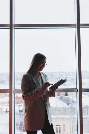 Confident slovenian business woman in elegant clothes checks notes in a notebook standing by the window in the office, a serious female manager reads information viewed on a notebookの写真素材