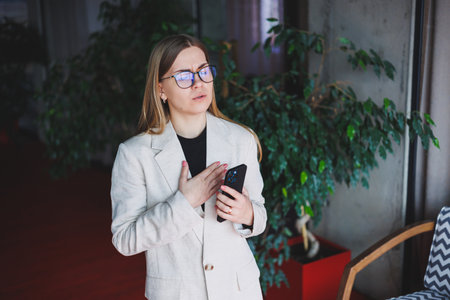 A beautiful young woman in a beige jacket and glasses read the news on a mobile phone and was...