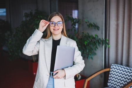 Portrait of a successful manager in the office smiling in during the working day, happy woman with laptop enjoying time in modern office. Blond-haired woman in a beige jacket and glassesの写真素材