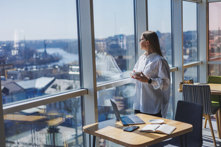 Portrait of a cheerful woman in classic glasses smiling in her free time in a cafe with coffee, positive jewish woman in a white shirt, desk with laptop, remote workの写真素材