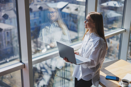 An elegant female businesswoman in glasses for vision correction, she holds a modern laptop in her hands, standing near a full-wall window in a modern office.の写真素材