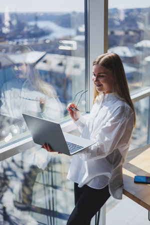 An elegant female businesswoman in glasses for vision correction, she holds a modern laptop in her hands, standing near a full-wall window in a modern office.の写真素材