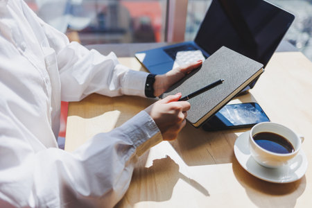 Notepad for entries in female hands on the background of a laptop close-up, a cup of coffee on a wooden table. Businessman's desk. Working process. selective focusの写真素材