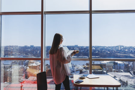 Thoughtful businesswoman with notepad, dressed in formal wear, standing near panoramic window in corporate company and thinking about trade investment, thoughtful businesswoman in glassesの写真素材