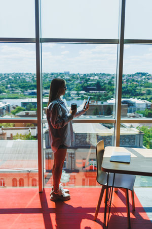 A young female freelancer in glasses is standing with coffee near a large window and looking at the...