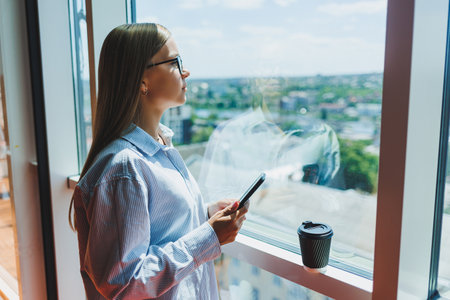 Attractive female blogger with a modern smartphone in her hands stands at the window, thoughtful...
