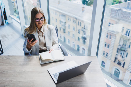 A fair-haired cheerful European woman in glasses in stylish casual clothes is sitting at a table with a laptop, doing paperwork and talking on the phone. Business lady at the workplace in the officeの写真素材
