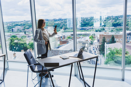 Cheerful young beautiful woman in glasses talking on a mobile phone and using a laptop with a smile while sitting at her workplace. Work in the officeの写真素材