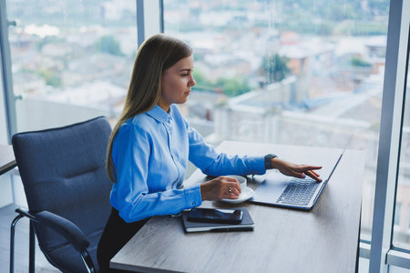Portrait of cheerful woman in classic glasses smiling in free time in cafe with coffee, positive european woman in blue shirt, desk with laptop, remote workの写真素材