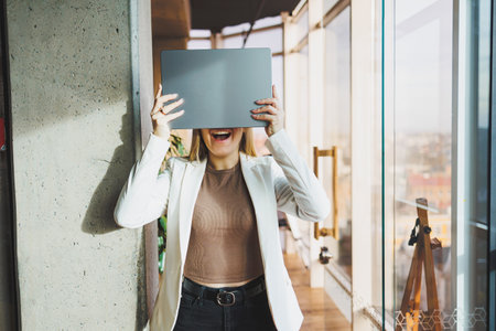 A young business woman with long blond hair in a white jacket and glasses stands with a laptop in...
