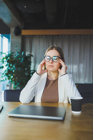 Tired young business woman sitting in the office at a wooden table behind a laptop. A young manager in glasses is tired of workの写真素材