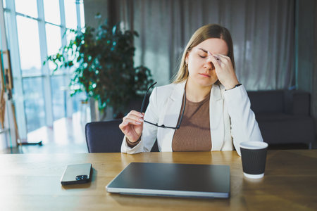 Tired young business woman sitting in the office at a wooden table behind a laptop. A young manager...