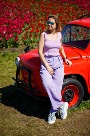 A girl model in casual clothes sits on a red car among tulip fields.の写真素材