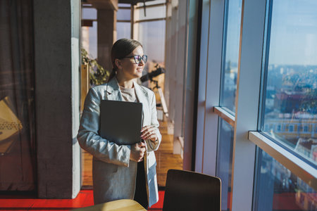 Cheerful young female freelancer in casual clothes standing at a wooden table with a netbook and writing notes while working on a project. Modern young business lady in the officeの写真素材