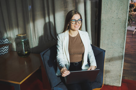 A young woman with long blond hair in stylish clothes is sitting in a chair and working on a laptop....