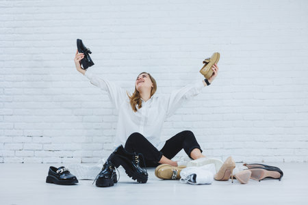Happy beautiful young woman with a smile in a fashionable outfit with a white shirt and black trousers sits on the floor among the shoes and chooses a new pair of shoes for herselfの写真素材