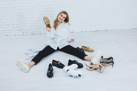 Stylish blonde choosing shoes while sitting on the floor. Side view of a lot of women's shoes on the...