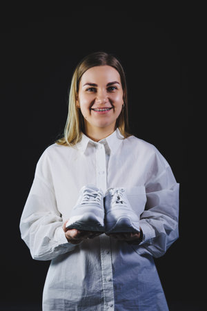 A fair-haired woman in a white shirt on a black background holds white sneakers made of genuine leather in her handsの写真素材