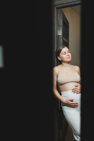 Cute young pregnant woman in a top and casual maternity clothes. Cute pregnant woman hugging her belly at homeの写真素材