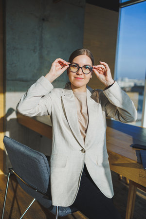 Work in a modern office. Positive young businesswoman in casual clothes and glasses sitting in the office with a mobile phone while working on a remote project using a laptopの写真素材