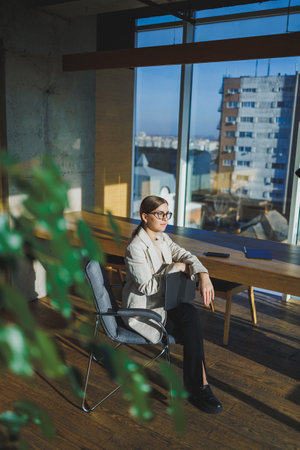 Smiling young businesswoman in casual clothes and glasses sitting in the office with a mobile phone while working on a remote project using a laptopの写真素材
