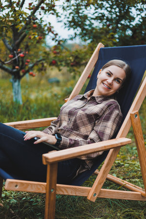 Beautiful woman in checkered shirt with laptop working outdoors in garden, home office concept. Remote work.の写真素材