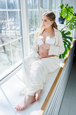Stylish pregnant young woman in a white suit near the window and touching her belly with a gentle smile. Awaiting birth. Care and motherhood. Love and tenderness.の写真素材