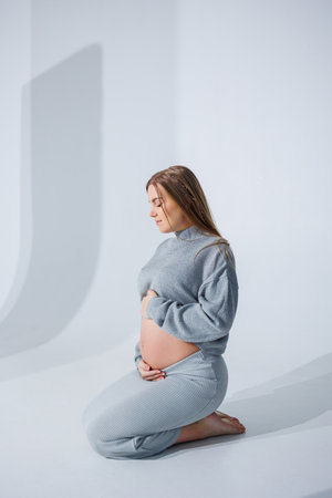 Cute young pregnant woman stroking her belly sitting on a white background. Stylish pregnant woman in skirt posing in studio and enjoying pregnancyの写真素材