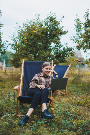 Stylish woman sitting on a chair near green plants in the garden and browsing the internet on a...