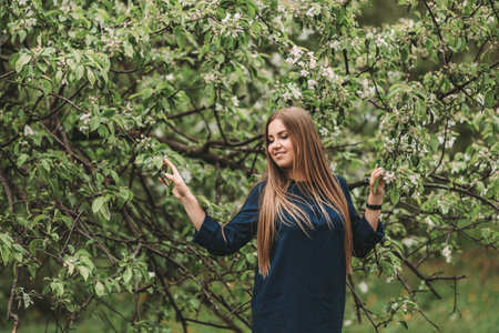 A beautiful young woman of Slavic appearance with long blond hair stands near a green tree with blossoms. She is wearing a blue dressの写真素材