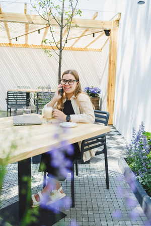 A woman manager sits on an outdoor terrace in a cafe and works online with coffee. Female freelancer works remotely online while sitting in a summer cafe. Remote workの写真素材