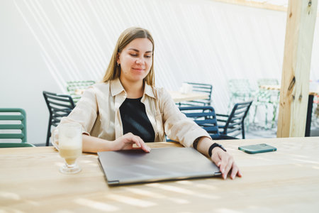 Young happy woman sitting at outdoor cafe table and talking on phone with cup of coffee, smiling...