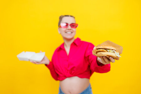 Hungry pregnant woman holding hamburger eating junk food posing on yellow background in studio. Woman enjoying a big hamburger. The concept of unhealthy eating and overeating during pregnancyの写真素材