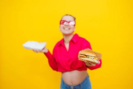 Hungry pregnant woman holding hamburger eating junk food posing on yellow background in studio. Woman enjoying a big hamburger. The concept of unhealthy eating and overeating during pregnancyの写真素材
