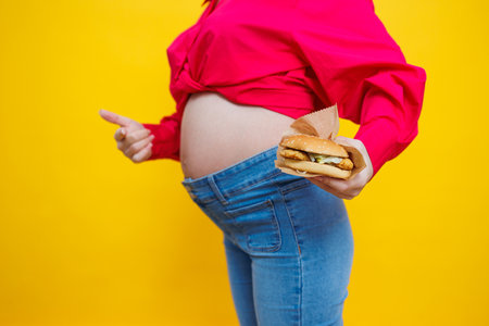 Cheerful pregnant woman in pink shirt holding hamburger over isolated yellow background with surprise and shocked facial expression. Harmful food during pregnancy. A pregnant woman eats fast foodの写真素材