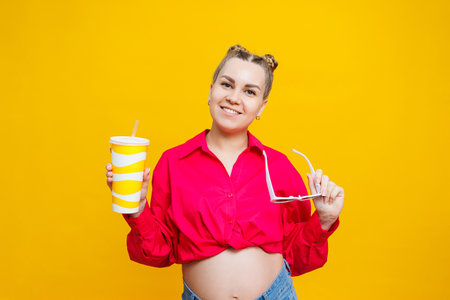 Cheerful pregnant woman in pink shirt holding tasty drink on isolated yellow background. A refreshing drink during pregnancy. A pregnant woman drinks water from a disposable glassの写真素材
