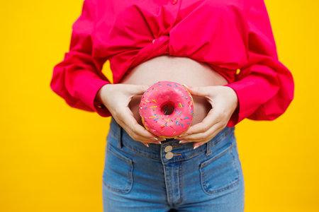 A beautiful pregnant woman holds a sweet donut in her hand on a yellow background. Expecting a...