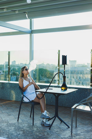 A young woman sits on an open terrace and smokes a hookah