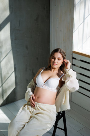 Stylish pregnant young woman in a white suit near the window and touching her belly with a gentle smile. Awaiting birth. Care and motherhood. Love and tenderness.の写真素材