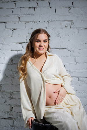 A stylish pregnant young woman in a white suit sits against a white brick wall and touches her belly with a gentle smile. Awaiting birth. Care and motherhood. Love and tenderness.の写真素材