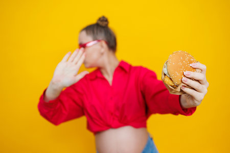 A cute pregnant woman in a pink shirt and pink glasses eats fast food. A pregnant woman on a yellow background with an appetizing burger in her hands. Harmful food for pregnant women.の写真素材