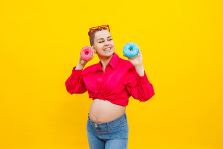 A bright pregnant woman in a pink shirt on a yellow background eats sweet donuts. Fresh sweet donuts in the hands of a pregnant womanの写真素材