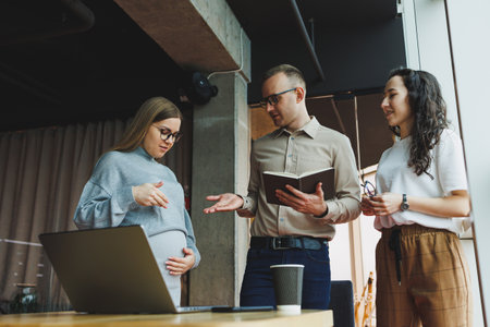 Modern pregnant woman working in office with colleagues. A young successful man helps a pregnant colleague at work. A beautiful pregnant woman works in the office. Modern pregnant womanの写真素材
