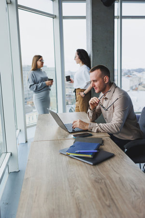 A young man works in the office on a laptop while sitting at the table against the background of...