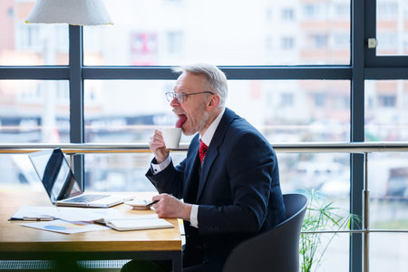 Male businessman sits with a cup of coffee and makes a new project with grimaces on his face.の写真素材