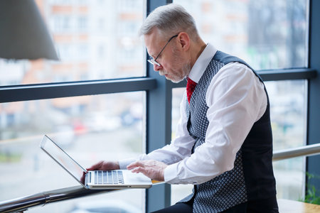 Adult male businessman, teacher, mentor working on a new project. Sits by a large window on the table. He looks at the laptop screen.の写真素材